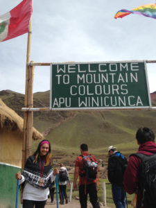 Leslie Peralta poses with a sign saying "Welcome to mountain colours APU Winicunca" 